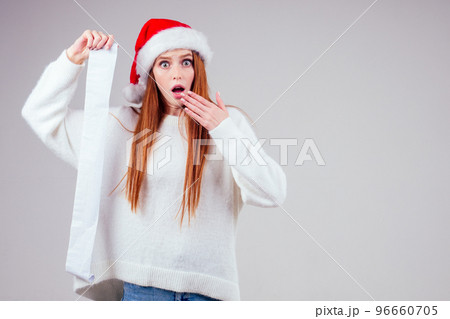 amazed and surprised redhaired ginger woman wearing red santa claus hat and knitted sweater holding long shopping receipt list in studio white background. big money xmas spending amazed and surprised redhaired ginger woman wearing red santa claus hat and knitted sweater holding long shopping receipt list in studio white background. big money xmas spending 96660705