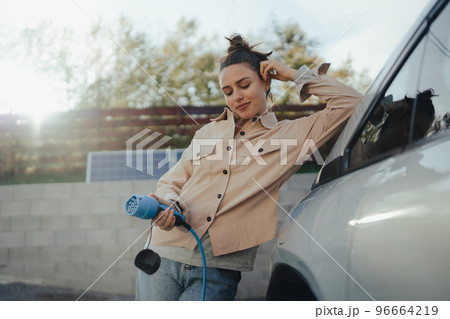 Young woman holding power supply cable from her electric car, prepared for charging it in home, sustainable and economic transportation concept. 96664219