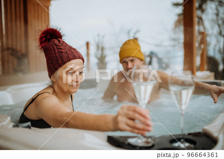 Senior couple in kintted cap enjoying together outdoor bathtub with glass of wine, at their terrace during cold winter day. Senior couple in kintted cap enjoying together outdoor bathtub with glass of wine, at their terrace during cold winter day. 96664361