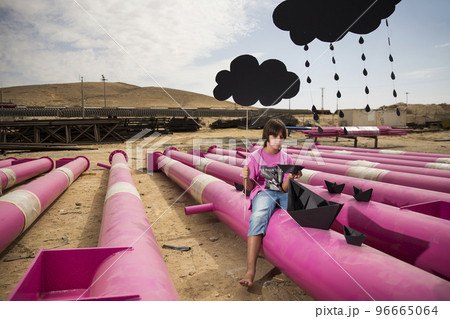 a cute kid in a pink t-shirt with a cat print plays among pink pipes with black clouds, rain, boats and paper planes against the blue sky in an industrial location. fashion style photoshoot 96665064