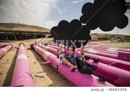 a cute kid in a pink t-shirt with a cat print plays among pink pipes with black clouds, rain, boats and paper planes against the blue sky in an industrial location. fashion style photoshoot a cute kid in a pink t-shirt with a cat print plays among pink pipes with black clouds, rain, boats and paper planes against the blue sky in an industrial location. fashion style photoshoot 96665098