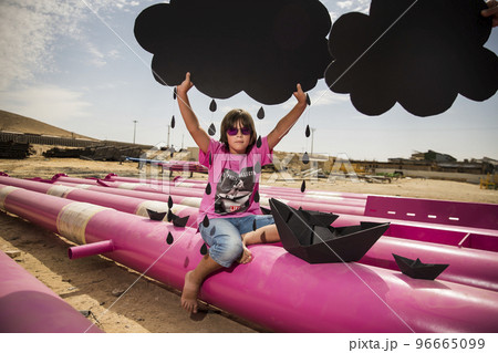 a cute kid in a pink t-shirt with a cat print plays among pink pipes with black clouds, rain, boats and paper planes against the blue sky in an industrial location. fashion style photoshoot a cute kid in a pink t-shirt with a cat print plays among pink pipes with black clouds, rain, boats and paper planes against the blue sky in an industrial location. fashion style photoshoot 96665099