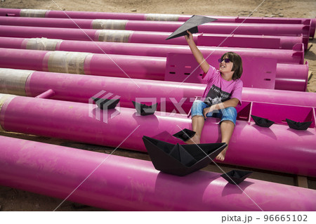 a cute kid in a pink t-shirt with a cat print plays among pink pipes with black clouds, rain, boats and paper planes against the blue sky in an industrial location. fashion style photoshoot 96665102