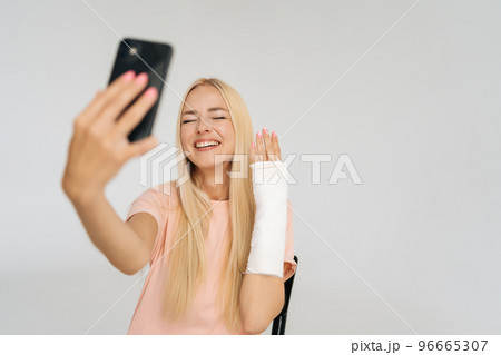 Studio portrait of positive young blonde woman with broken arm wrapped in plaster bandage talking smartphone making video call, taking selfie picture, standing on white isolated background. Studio portrait of positive young blonde woman with broken arm wrapped in plaster bandage talking smartphone making video call, taking selfie picture, standing on white isolated background. 96665307