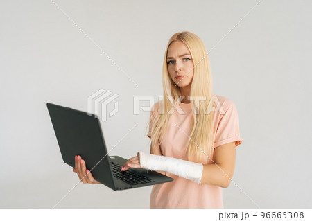 Portrait of serious blonde young woman with broken arm wrapped in plaster bandage holding laptop, looking at camera, standing on white isolated background. Concept of insurance and healthcare Portrait of serious blonde young woman with broken arm wrapped in plaster bandage holding laptop, looking at camera, standing on white isolated background. Concept of insurance and healthcare 96665308
