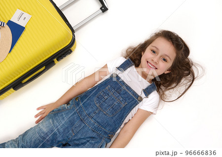 Charming European little child, adorable baby girl passenger wearing blue denim overalls, lies on white background near her boarding pass on yellow suitcase, smiles a beautiful toothy smile to camera Charming European little child, adorable baby girl passenger wearing blue denim overalls, lies on white background near her boarding pass on yellow suitcase, smiles a beautiful toothy smile to camera 96666836
