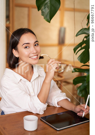 Beautiful asian girl sitting in cafe with cup of coffee, drawing on digital tablet with graphic pen, doddling and making scatches Beautiful asian girl sitting in cafe with cup of coffee, drawing on digital tablet with graphic pen, doddling and making scatches 96667791