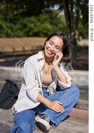Smiling asian girl relaxing in park, talking on smartphone, having a mobile call while resting outdoors 96667805