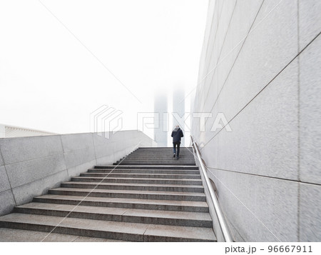 Man is going upstairs in underground pedestrian crossing. Tops of skyscrapers are in fog. Loneliness in big city. Urban architecture and lonely man. Monochrome background. 96667911