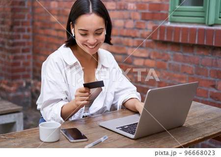 Smiling asian woman with laptop and wireless earphones, paying with credit card, buying online, sitting with cup of coffee Smiling asian woman with laptop and wireless earphones, paying with credit card, buying online, sitting with cup of coffee 96668023