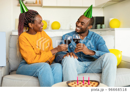 Loving black spouses clinking glasses of wine, celebrating birthday at home, sitting on sofa and smiling at each other 96668518
