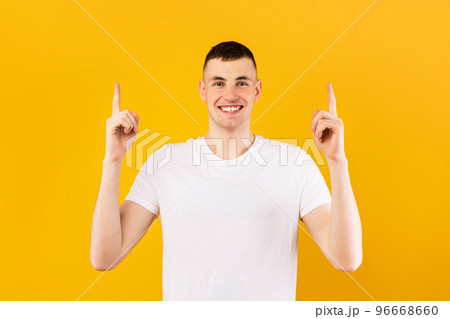 Happy caucasian man in white t-shirt pointing up at empty space and smiling at camera, standing over yellow background Happy caucasian man in white t-shirt pointing up at empty space and smiling at camera, standing over yellow background 96668660