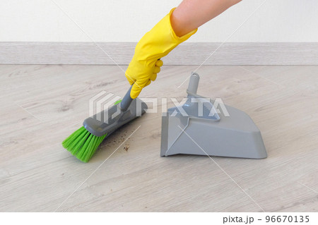 Cleaning concept. Female hand in yellow rubber glove sweeping wooden floor with green brush and gray scoop indoor. 96670135
