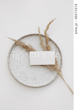 Blank textured paper business card mockup. Ceramic plate isolated on white table background. Boho stationery with dry grass. Branding template. Minimal composition. Vertical flat lay, top view. 96673978