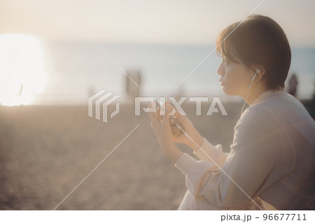 夕日をバックにスマホで音楽を聴く若い女性の写真 96677711