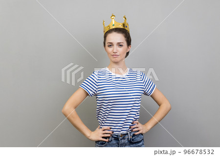Portrait of woman wearing striped T-shirt and golden crown, looking at camera with confident facial expression, keeps hands on hips. Indoor studio shot isolated on gray background. 96678532