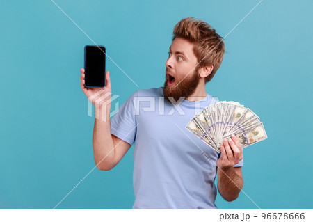 Portrait of surprised amazed handsome bearded man holding dollar banknotes and looking at mobile phone with empty screen, online betting, winning. Indoor studio shot isolated on blue background. 96678666