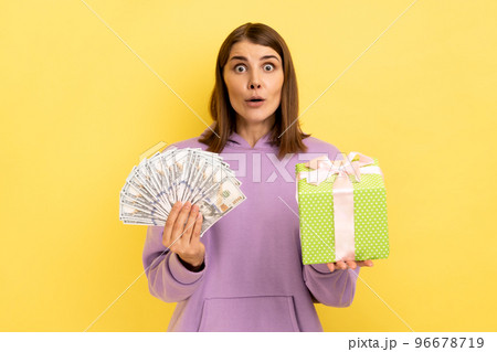 Portrait of excited amazed woman holding wrapped present box and doolars banknotes, looking at camera with open mouth, wearing purple hoodie. Indoor studio shot isolated on yellow background. Portrait of excited amazed woman holding wrapped present box and doolars banknotes, looking at camera with open mouth, wearing purple hoodie. Indoor studio shot isolated on yellow background. 96678719