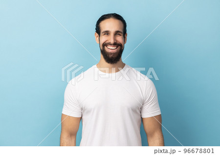 Portrait of bearded handsome man wearing white T-shirt standing looking at camera with satisfied face and smiling, expressing happiness. Indoor studio shot isolated on blue background. 96678801
