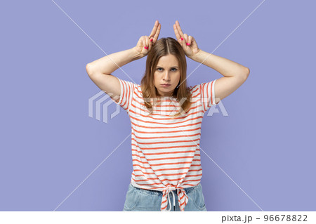 Portrait of angry bossy blond woman wearing striped T-shirt standing and showing horns, being anger and aggressive, looking at camera. Indoor studio shot isolated on purple background. Portrait of angry bossy blond woman wearing striped T-shirt standing and showing horns, being anger and aggressive, looking at camera. Indoor studio shot isolated on purple background. 96678822