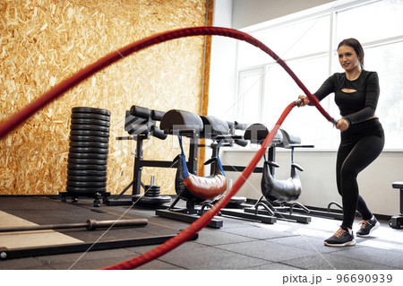 Attractive young slim and fit sportswoman working out in the gym for functional training doing crossfit exercises with battle ropes 96690939