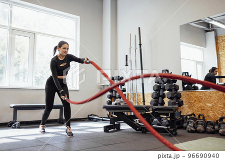 Attractive young slim and fit sportswoman working out in the gym for functional training doing crossfit exercises with battle ropes 96690940