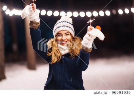 Cheerful young woman holding a sparkler in hand in the winter forest. Happy cute girl posing with sparkler. Cheerful young woman holding a sparkler in hand in the winter forest. Happy cute girl posing with sparkler. 96691930