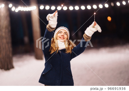 Cheerful young woman holding a sparkler in hand in the winter forest. Happy cute girl posing with sparkler. Cheerful young woman holding a sparkler in hand in the winter forest. Happy cute girl posing with sparkler. 96691936