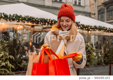 Attractive blonde young woman shopping, holding shopping red bags and gift boxes with red Christmas hat, smiling while standing in front of shop window. Outdoors.  96696165