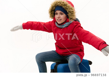 Teenage boy in red parka with fur hood, sits on blue suitcase, stretches his arms to the sides, imitating airplane during flight, isolated on white background. Winter travel concept. Holiday. Vacation Teenage boy in red parka with fur hood, sits on blue suitcase, stretches his arms to the sides, imitating airplane during flight, isolated on white background. Winter travel concept. Holiday. Vacation 96697138