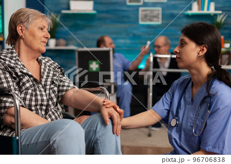 Female nurse supporting elderly woman with disability in nursing home waiting room. Male nurse taking temperature of senior patient with infrared thermometer, seated on couch in background. 96706838