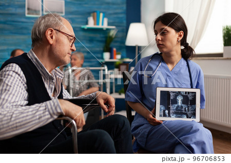 Nurse showing an Xray on a tablet to a patient with disability in a nursing home. Retired man sitting in a wheelchair looking at radiograph and listening to directions for recovery. 96706853