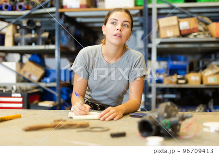 Young woman storehouse worker standing at workbench and making notes Young woman storehouse worker standing at workbench and making notes 96709413
