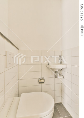 Interior of narrow restroom with sink and wall hung toilet with white walls and tile floor Interior of narrow restroom with sink and wall hung toilet with white walls and tile floor 96712903