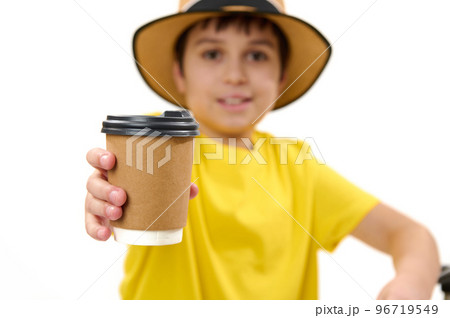 Selective focus on a disposable cardboard cup with takeaway hot drink in the hands of a blurred teenage boy in yellow t-shirt and straw hat, handing paper mug on camera, isolated over white background Selective focus on a disposable cardboard cup with takeaway hot drink in the hands of a blurred teenage boy in yellow t-shirt and straw hat, handing paper mug on camera, isolated over white background 96719549