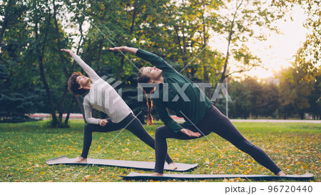 Two pretty women are doing yoga outdoors in city park on mats practising asanas and breathing fresh air. Individual practice, professional instructor and nature concept. 96720440