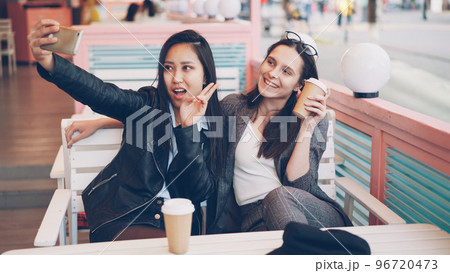 Carefree girls are taking selfie with take out drinks sitting in cafe together and using smartphone. Young women are posing, clinking glasses and showing hand gestures v-sign. 96720473