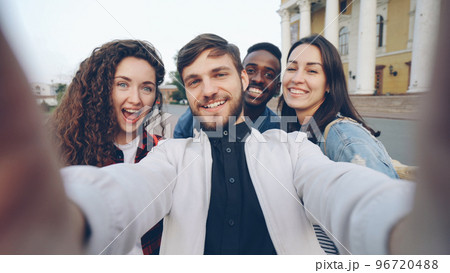 Point of view shot of multiracial group of tourists taking selfie in city center holding gadget camera and posing together with hand gestures expressing positive emotions. 96720488