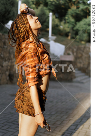 a girl with a dreadlocked hairstyle poses in summer outdoor, bright sunlight, dressed in a plaid shirt and shorts 96722031