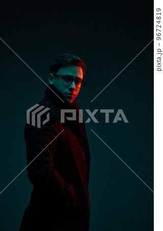 Studio shot of serious young 30s years old man in classic style clothes posing isolated over dark background in neon light. Fashion, style, emotions concept Studio shot of serious young 30s years old man in classic style clothes posing isolated over dark background in neon light. Fashion, style, emotions concept 96724819