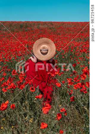 Young woman stands with her back in a long red dress and hat, posing on a large field of red poppies 96725458
