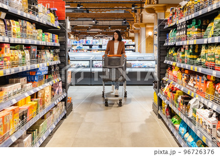 a woman with a cart walks between rows of shelves in a grocery store 96726376