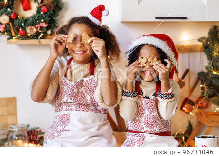 Happy ethnic children on Christmas eve,   girl and boy covers eyes with cookies gingerbreads that they baked and laughs  in cozy kitchen 96731046