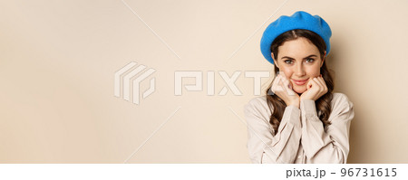 Close up portrait of feminine young woman in trendy french hat, posing cute and romantic, making silly coquettish face and tender smile, beige background 96731615