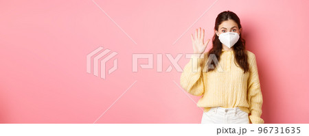 Covid-19, lockdown and pandemic concept. Young woman wearing face mask during quarantine, saying hello, waving raised hand to greet person from distance, pink background 96731635