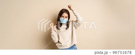 Covid-19, pandemic and quarantine concept. Enthusiastic girl dancing in face medical mask, celebrating, standing over beige background Covid-19, pandemic and quarantine concept. Enthusiastic girl dancing in face medical mask, celebrating, standing over beige background 96731678