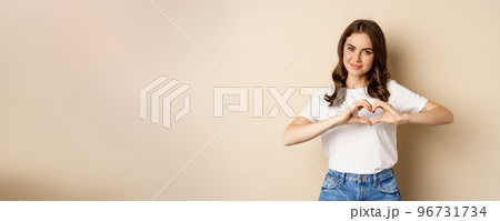 Lovely woman showing heart, love sign and smiling, standing over beige background in white t-shirt, beige background Lovely woman showing heart, love sign and smiling, standing over beige background in white t-shirt, beige background 96731734
