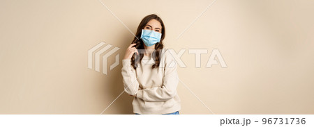 Covid-19, pandemic and quarantine concept. Beautiful modern woman in medical face mask, cross arms on chest, looking confident, beige background Covid-19, pandemic and quarantine concept. Beautiful modern woman in medical face mask, cross arms on chest, looking confident, beige background 96731736