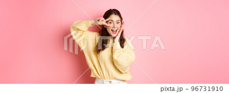 Attractive female model, gasping amazed, showing v-sign over eye, smiling happy, standing against pink background 96731910