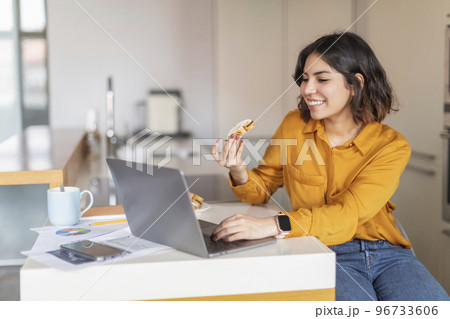 Smiling Young Arab Woman Eating Snack And Using Laptop In Kitchen Smiling Young Arab Woman Eating Snack And Using Laptop In Kitchen 96733606